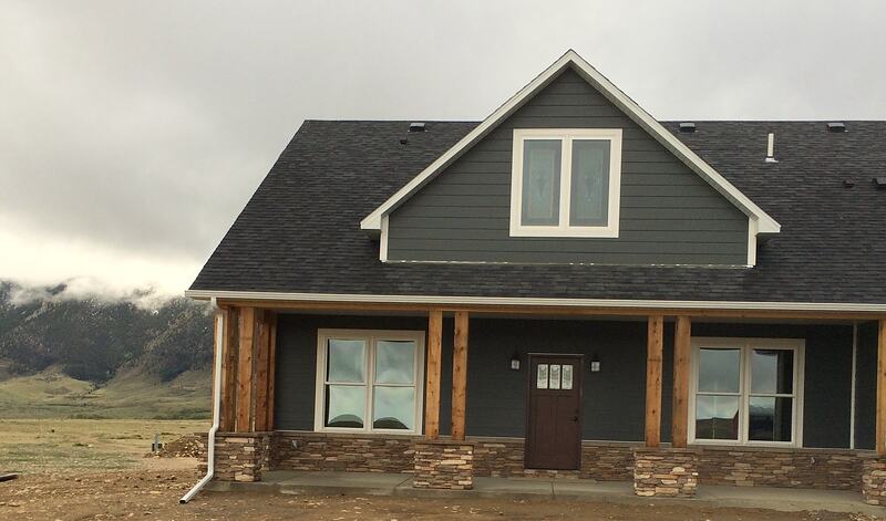 Front of the home with dark gray siding, a gable window above a covered porch on wood posts and stacked stone bases, and mountains behind