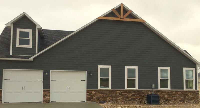 Side of the home with a two-car garage with white carriage-style doors, dark gray siding over a stacked stone wainscot, and a wood truss detail in the gable