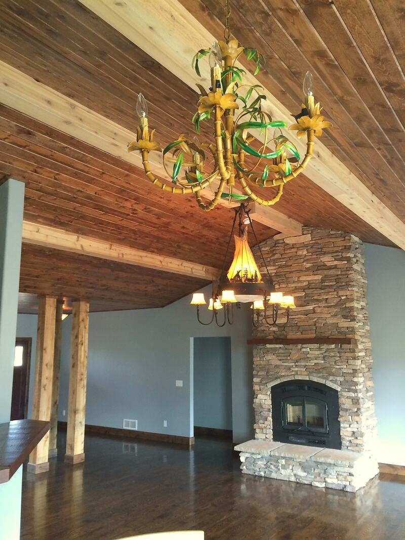 Great room with two chandeliers under a vaulted wood plank ceiling with light wood beams, rough wood posts, dark hardwood floors and the stacked stone fireplace