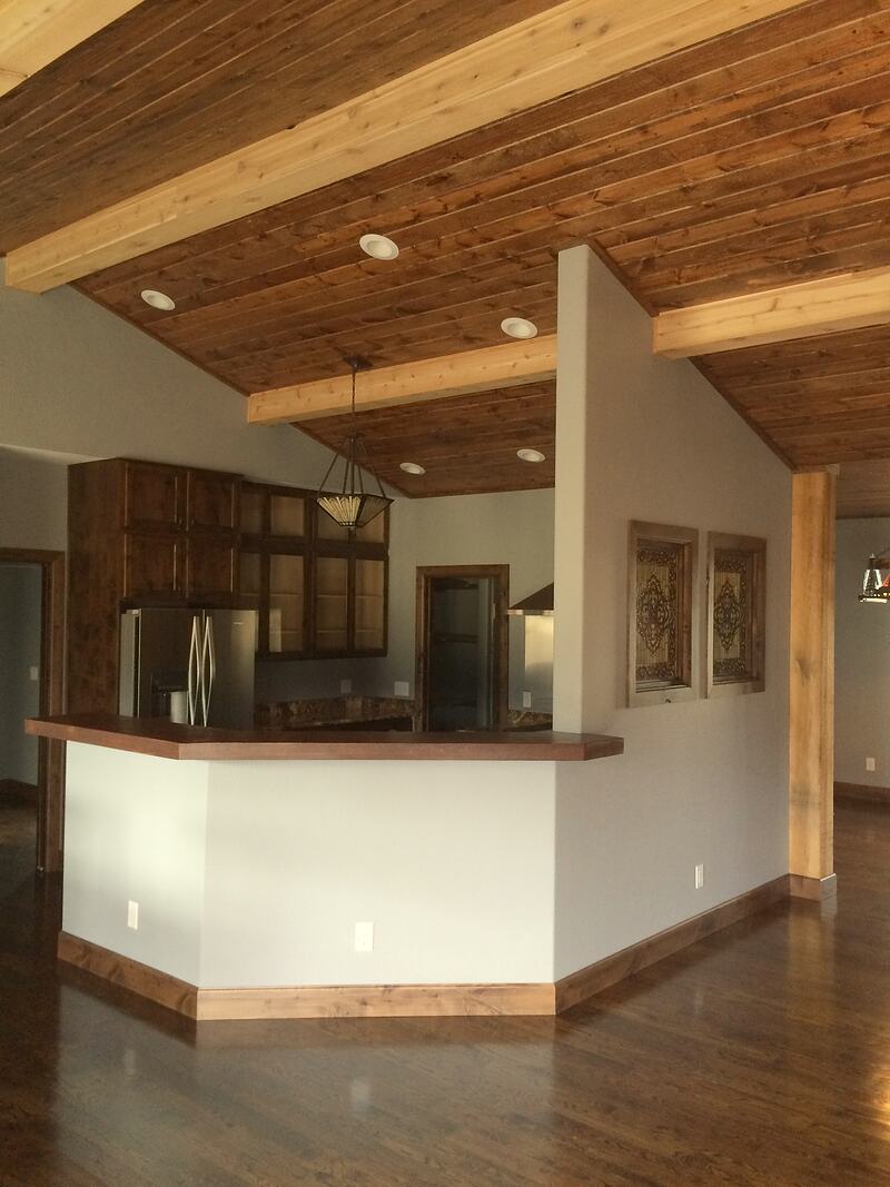 Kitchen behind an angled bar with a dark wood top, with dark wood cabinets, a stainless steel refrigerator, recessed lights and a pendant light under the wood ceiling